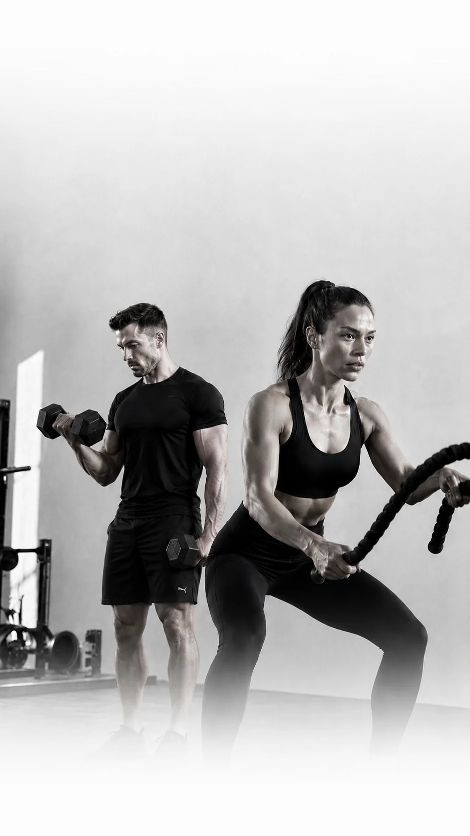 Two athletes training with dumbbells and a battle rope in a bright gym.